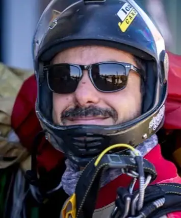 Tandempilot Daniel mit seinem Tandemschirm über der Schulter und Helm lächelt in die Kamera.